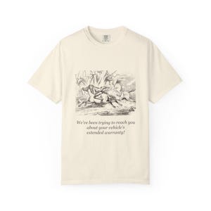 Pode incluir: Uma t-shirt de manga curta na cor creme com uma ilustração de linhas a preto e branco de uma pessoa a montar um cavalo, com o texto "We've been trying to reach you about your vehicle's extended warranty!" impresso abaixo.