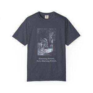 May include: A dark charcoal gray t-shirt featuring a rectangular black and white illustration of a person sitting in a dimly lit room. Below the graphic, the text reads: Mourning Person, Not a Morning Person. The shirt has a crew neckline.