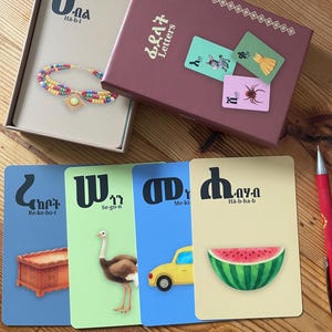 May include: A set of educational flashcards in a maroon box with the text Letters. The cards feature Amharic characters, phonetic pronunciations, and illustrations including a beaded necklace, a chest, an ostrich, a car, and a watermelon slice.