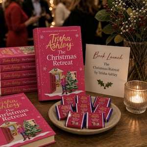 May include: A stack of pink books titled The Christmas Retreat by Trisha Ashley, featuring a festive illustration of a person by a Christmas tree. A plate holds small chocolate bars with matching covers, next to a sign reading Book Launch.