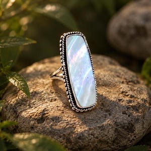 May include: A silver ring featuring a long, rectangular, iridescent mother-of-pearl inlay. The stone is framed by a twisted rope-style metal border, and the band consists of a split shank design that tapers towards the back of the ring.