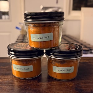 Puede incluir: Tres frascos pequeños de vidrio transparente con tapas de metal negro, llenos de polvo de raíz de cúrcuma de color naranja vibrante. Cada frasco tiene una etiqueta rectangular azul claro con el texto Turmeric Root impreso en negro.