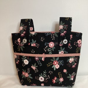 Peut inclure: Un sac fourre-tout noir avec un motif floral de fleurs roses, rouges et blanches. Le sac a deux poignées et une bordure rose. Des boutons argentés fixent les poignées au sac.