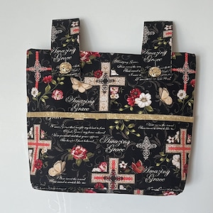 May include: Black fabric tote bag with a floral and cross pattern. The bag features the words "Amazing Grace" and gold-colored accents. The design includes red and white flowers, butterflies, and decorative crosses.
