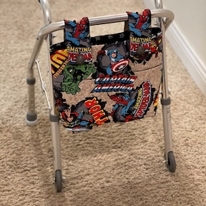 May include: A silver walker with a fabric bag attached to the handles. The bag is made of a beige fabric with a comic book pattern featuring superheroes like Captain America, Spiderman, Hulk, and Thor. The text on the bag reads "Amazing Spiderman" and "Captain America".