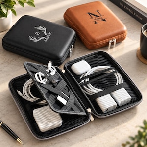 Peut inclure: Deux étuis de rangement rectangulaires zippés pour accessoires électroniques en cuir noir et marron clair. L'étui noir présente un logo en forme de bois de cerf blanc et le nom BAKER. L'étui marron affiche un grand N et le nom Nelson.