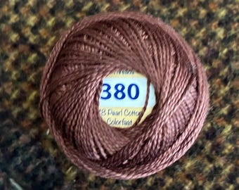 May include: A round ball of brown pearl cotton embroidery thread. The center label is white with yellow accents and displays the number 380 in blue text, along with the words 8 Pearl Cotton and Colorfast printed in black.