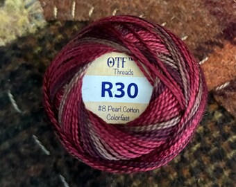 May include: A round ball of variegated embroidery thread featuring shades of deep red, burgundy, and brown. A central label displays the text OTF Threads, R30, and #8 Pearl Cotton Colorfast in blue and black ink on a cream-colored background.