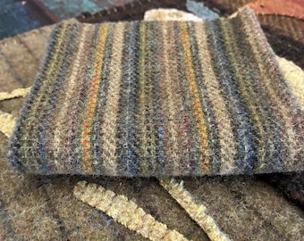 May include: A folded piece of thick, textured wool fabric featuring vertical stripes in shades of olive green, brown, blue, and yellow. The material has a visible woven construction with a fuzzy, felted surface texture throughout the pattern.