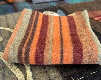 May include: A folded piece of thick, textured wool fabric featuring vertical stripes in shades of burnt orange, deep maroon, brown, and muted sage green. The material has a fuzzy, felted appearance with a distinct woven pattern throughout the textile.