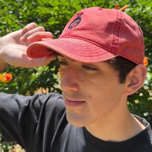 May include: A red baseball cap with a distressed, washed-out finish. The cap features a black embroidered circular logo on the front panel, which depicts a stylized insect silhouette. The design includes a curved brim and stitched paneling.