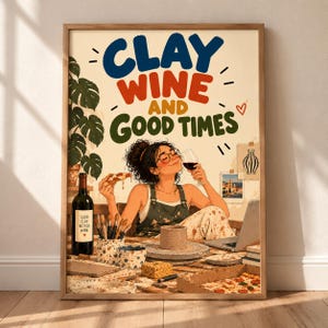 May include: A framed wall art print featuring the text CLAY WINE AND GOOD TIMES in blue, red, and green. The illustration depicts a person with glasses holding a slice of pizza and a wine glass, sitting at a pottery table with a laptop, brushes, and a wine bottle labeled GOOD CLAY BETTER WINE.