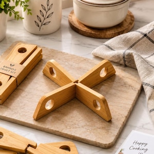 May include: A set of light brown bamboo trivets featuring a cross-shaped design with circular cutouts at each end. The set includes one folded stack secured with a paper band labeled BAMBOO TRIVET SET, one assembled cross-shaped trivet, and two loose pieces.