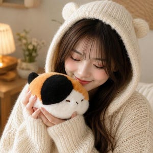 May include: A cream-coloured knit hooded jumper featuring small rounded ears on the hood, paired with a small, plush, tri-coloured calico cat toy with black, orange, and white fur, held close to the chest in a soft, textured fabric design.