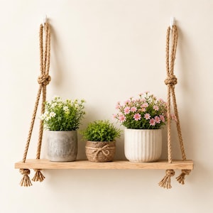 Puede incluir: Una repisa rectangular de madera suspendida por gruesas cuerdas de yute color canela, con tres plantas en maceta. La maceta izquierda es gris, la central es de mimbre marrón y la derecha es de cerámica blanca acanalada con flores rosas.