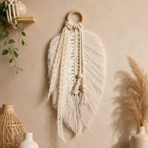 May include: A cream-colored macrame wall hanging shaped like a large feather, suspended from a circular wooden ring. The piece features intricate knotted cord, a delicate lace ribbon trim, and two smooth wooden beads hanging from the center.