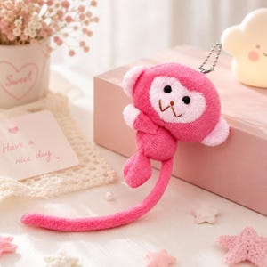 May include: A plush monkey keyring featuring bright pink fur, a light pink face, and a long, slender tail. The monkey has black embroidered eyes and a small brown mouth. A silver metal ball chain is attached to the top of the monkey's head.