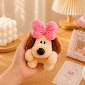 May include: A small plush dog toy with cream-coloured fur, brown ears, and a large, textured pink bow on its head. The dog features a smooth, round black nose and two white eyes with black pupils, crafted from a soft, fuzzy material.