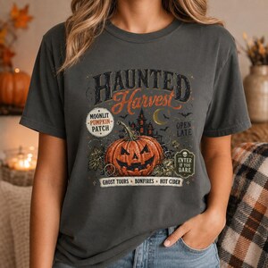 May include: A charcoal gray t-shirt featuring a graphic print with the text Haunted Harvest. The design includes a large orange jack-o'-lantern, a dark silhouette of a haunted house, a crescent moon, bats, and text reading Moonlit Pumpkin Patch, Open Late, and Enter If You Dare.