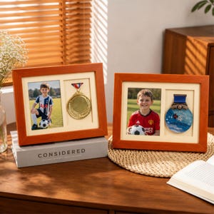 May include: Two rectangular wooden frames with a warm brown finish, each featuring a dual-aperture mat. One side holds a photograph of a child in a soccer uniform, while the other side displays a sports medal with a ribbon, preserved behind glass.