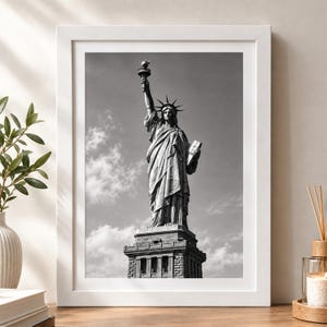 Könnte beinhalten: Ein schwarz-weiß gerahmter Druck der Freiheitsstatue auf ihrem Sockel, die eine Fackel in der einen und eine Tafel in der anderen Hand hält, vor einem Himmel mit vereinzelten Wolken, eingefasst in einen schlichten weißen Rahmen.