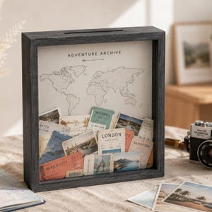 May include: A dark gray wooden shadow box frame with a slot on top, featuring a world map background labeled ADVENTURE ARCHIVE. The interior is filled with various travel tickets, postcards, and photographs from locations like London, Mexico City, and Barcelona.