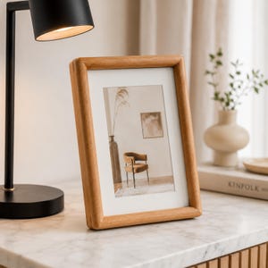 Puede incluir: Un marco de fotos rectangular hecho de madera de tono claro con un acabado liso. El marco contiene una impresión que representa una escena interior con una silla, un jarrón y arte de pared. Se mantiene en pie junto a una lámpara de escritorio negra.