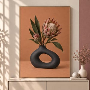 Puede incluir: Una impresión de arte mural enmarcada que muestra un jarrón gris oscuro asimétrico con un centro hueco, que contiene dos flores de protea con pétalos blanco rosáceos y hojas verdes largas, sobre un fondo sólido de color terracota.