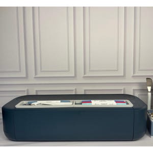 May include: A dark blue electronic cutting machine with a smooth, rounded rectangular exterior. The top surface features a recessed light gray storage tray containing various pens, markers, and a small metal tool for crafting projects.