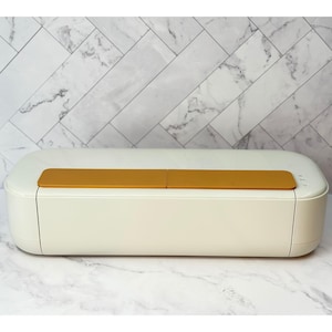 May include: A cream-colored electronic cutting machine with a smooth, rounded rectangular shape. The top features two rectangular mustard-yellow panels. On the right side, there are small control buttons and indicator lights on the casing.