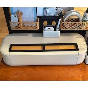 May include: A white electronic cutting machine with a black top panel featuring two rectangular inserts. Above the machine, a black organizer holds two tan honeycomb-patterned bins, a white pen holder with a black marker, and various craft supplies.