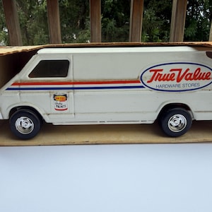 May include: A white Ertl Toys die-cast metal van in its original cardboard box. The van features red and blue side stripes, a True Value Hardware Stores logo, and a small graphic of a paint can labeled Top Test Quality Paints on the side panel.