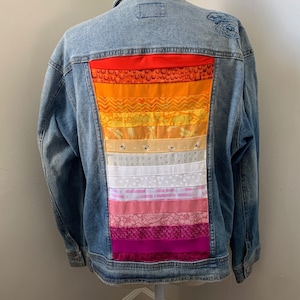 May include: A blue denim jacket with a multi-colored patchwork design on the back. The patchwork is made up of different fabrics in shades of red, orange, yellow, white, pink, and purple. The design resembles a pride flag.