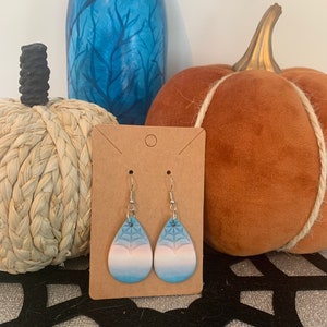 Puede incluir: Un par de pendientes colgantes con un diseño degradado azul, rosa y blanco. Los pendientes presentan un patrón de telaraña y ganchos plateados.