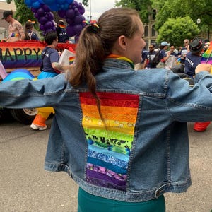 May include: A person wearing a blue denim jacket with a rainbow-colored patchwork design on the back. The person is wearing green pants and has long brown hair.