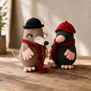 May include: Two crocheted mole dolls. The left mole is light brown with a black hat, round wire-rimmed glasses, and a dark red scarf. The right mole is dark gray with a red hat and a matching dark red scarf. Both have pink paws.