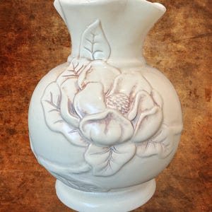 May include: A cream-colored ceramic vase featuring a prominent embossed floral design on the front. The vase has a rounded body, a flared neck, and a wide base. The flower relief displays subtle brownish shading within the petals and leaves.