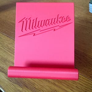 Op de afbeelding: Een felrode, schuine telefoon- of tablethouder met het Milwaukee-logo in reliëf op het voorvlak. De houder heeft een plat achterpaneel en een horizontale cilindrische rand aan de onderkant om een apparaat rechtop te ondersteunen.
