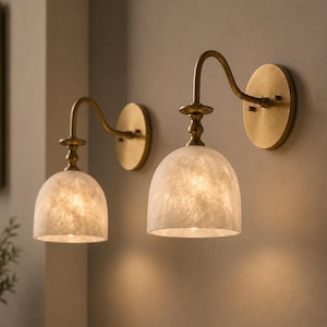 May include: Two wall-mounted light fixtures featuring curved brass arms and circular backplates. Each lamp has a dome-shaped shade made of translucent, marbled white material that emits a soft, warm glow, illuminating the wall surface.