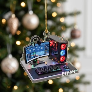 May include: A flat acrylic Christmas ornament shaped like a gaming desk setup with a curved monitor, a tower PC with red and blue glowing fans, a keyboard, a mouse, and headphones. The desk surface features the name ETHAN printed in white text.