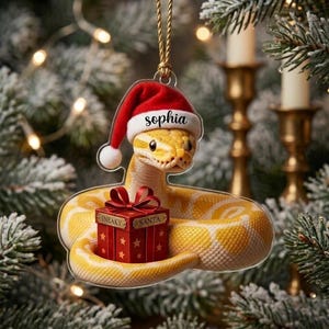 May include: A clear acrylic hanging ornament featuring a yellow snake wearing a red and white Santa hat with the name Sophia printed on the brim. The snake is coiled around a red gift box with a bow, labeled with the words SNEAKY and SANTA.
