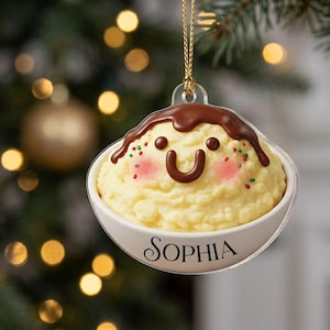 May include: A round, clear acrylic Christmas ornament featuring a 3D-style illustration of a bowl of rice pudding. The pudding has a smiling face, pink cheeks, chocolate sauce topping, and colorful sprinkles. The name SOPHIA is printed in black.