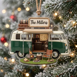 May include: A flat, decorative ornament shaped like a green and white camper van with an open side revealing a detailed interior. The roof features a sign reading The Millers, luggage, and a lantern. Outside, a small stove and chair sit on grass.