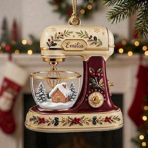 May include: A cream and deep red stand mixer ornament featuring gold accents, holly leaf and berry motifs, and the name Emilia printed on the front. The clear glass mixing bowl contains a miniature snow-covered gingerbread house and pine trees.