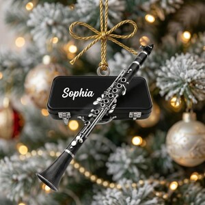 May include: A black clarinet ornament featuring silver-toned keys and a black rectangular instrument case with the name Sophia printed in white script. The ornament is suspended by a gold-colored cord tied in a decorative bow at the top.