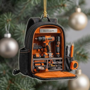May include: A flat, hanging ornament shaped like an open black and orange backpack. The interior displays a drill, flashlight, tape measure, and tools. A silver nameplate at the top is engraved with the name William. It hangs from a gold cord.