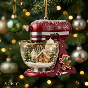 May include: A red stand mixer Christmas ornament featuring a clear glass bowl filled with a miniature gingerbread house, two pine trees, and snow. The mixer is decorated with white snowflakes, a gingerbread man, and the name Sophia in white script.