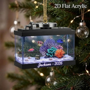May include: A 2D flat acrylic Christmas tree ornament shaped like a rectangular aquarium with a black frame. Inside are colorful coral, a clownfish, and a blue tang fish on white sand. The text Jackson - 2026 is printed on the front black base.
