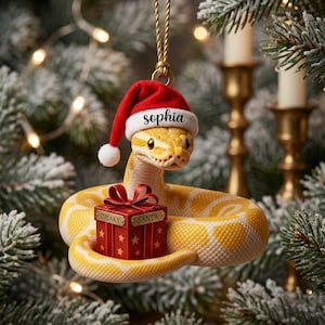 May include: A yellow and white snake Christmas ornament wearing a red and white Santa hat with the name Sophia printed on the brim. The snake is coiled around a red gift box with a gold bow and the text Sneaky Santa printed on the front.
