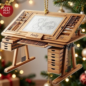 May include: A flat, 2D wooden drafting table ornament featuring a grid surface with a bicycle sketch, integrated storage compartments, and a drawer unit. The name Andy is engraved on the front edge. A gold cord is attached for hanging.